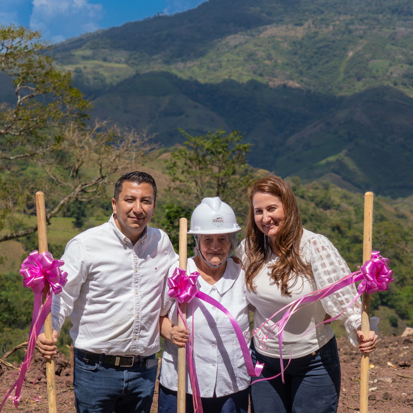 Tres personas sonríen con palas decoradas con cintas rosadas, de pie en el terreno de Vista Hermosa con las montañas de Naranjito al fondo